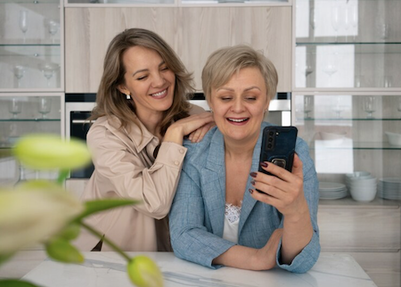 A middle aged woman with her hands on her older mother's shoulders while they are looking at something on the smartphone together